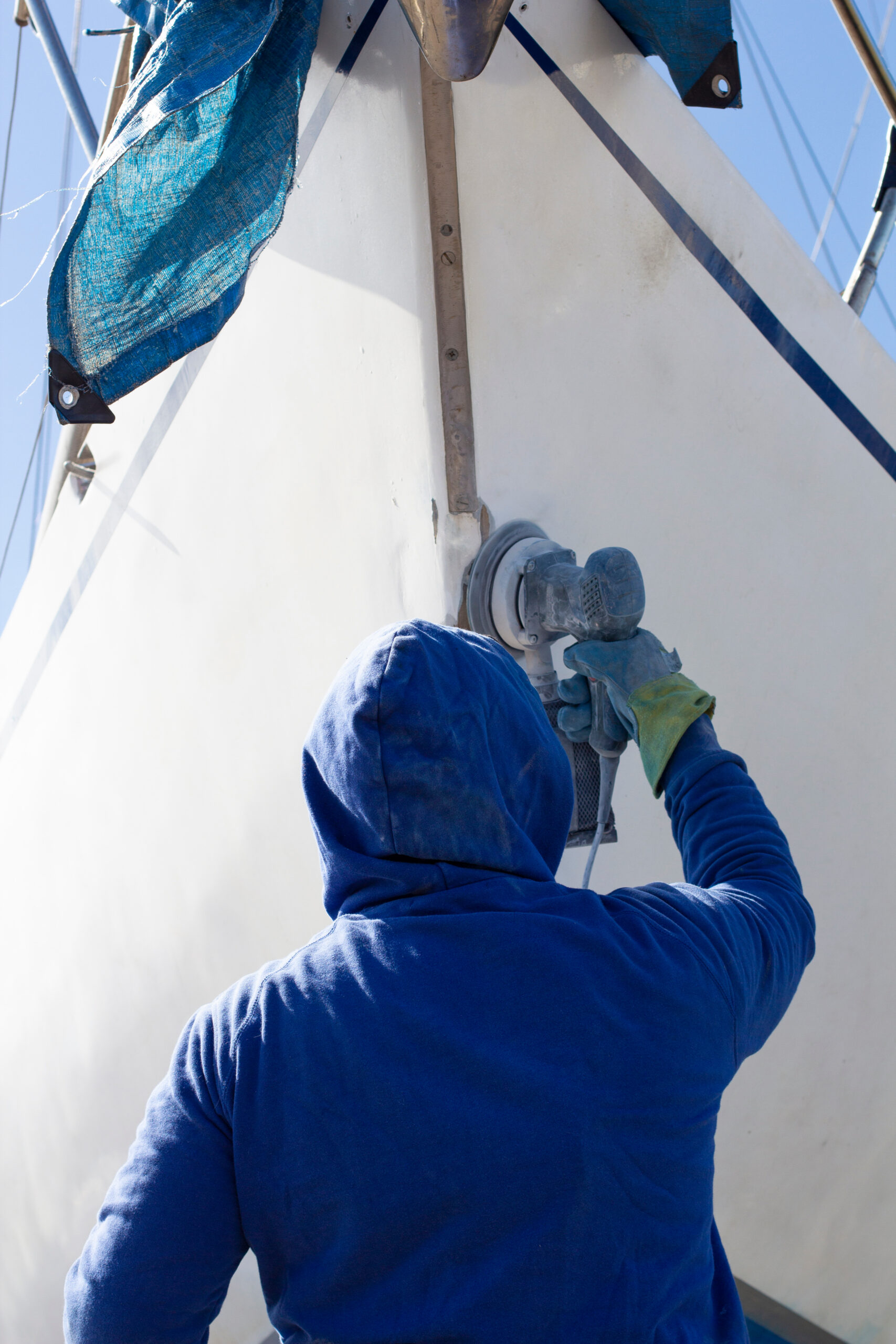 A man in a blue jacket polishes his yacht with a grinder before painting.